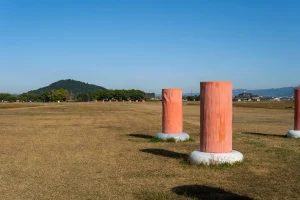 広い草地に立つ朱色の復元柱と、そのむこうに見える耳成山